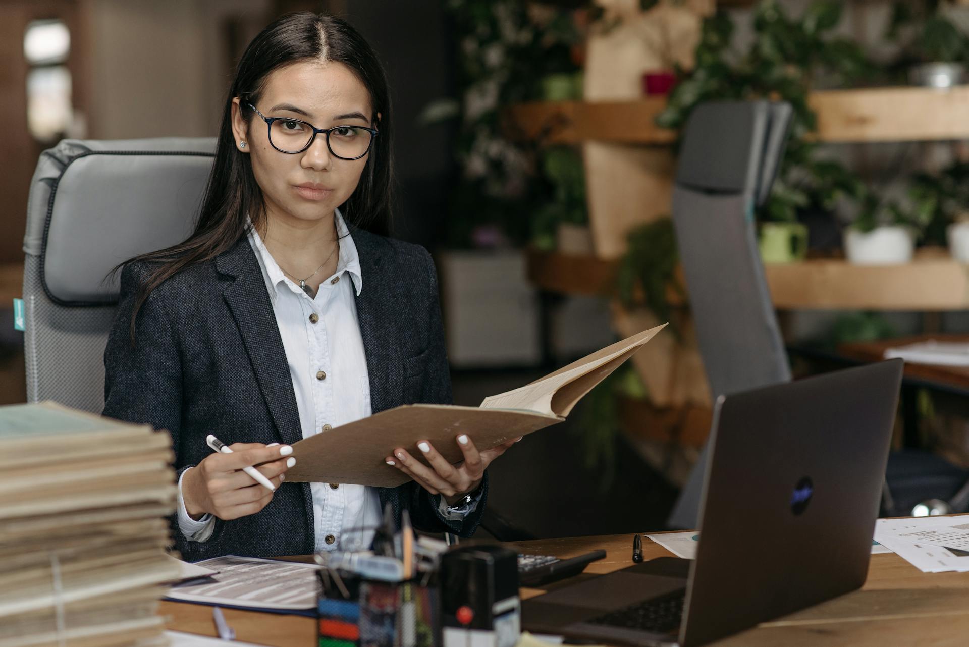 Confident businesswoman in professional attire reviewing documents at her desk in a modern office setting.