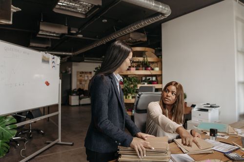 Free Two professional women discussing work in a modern office setting with documents and a whiteboard. Stock Photo