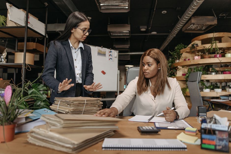 Dos mujeres de negocios en traje corporativo trabajando juntas en un entorno de oficina moderno con documentos.
