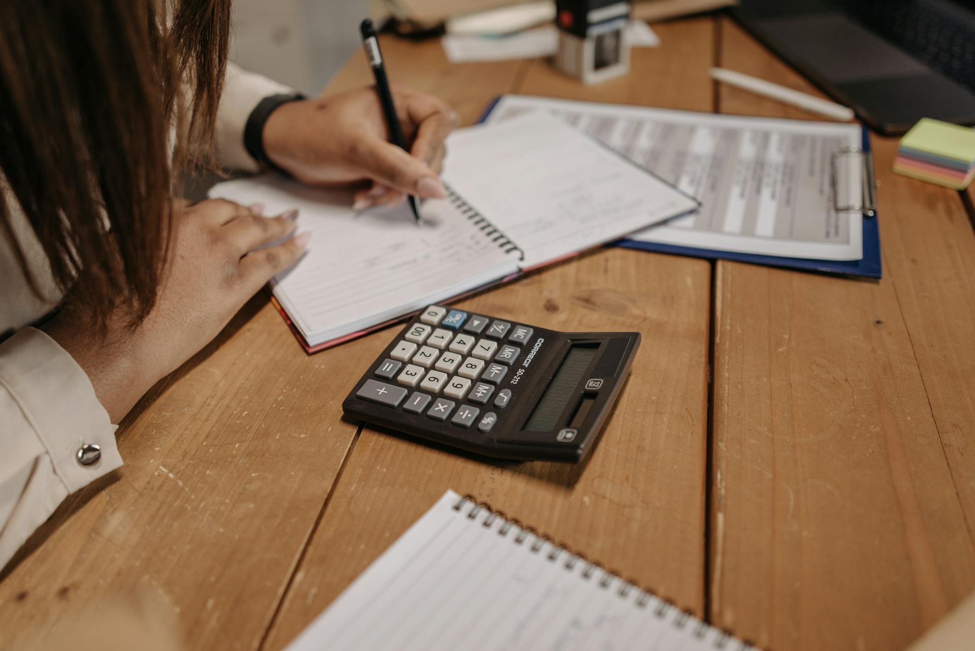 Woman writing notes with a calculator on a wooden desk, focusing on work and accounting.