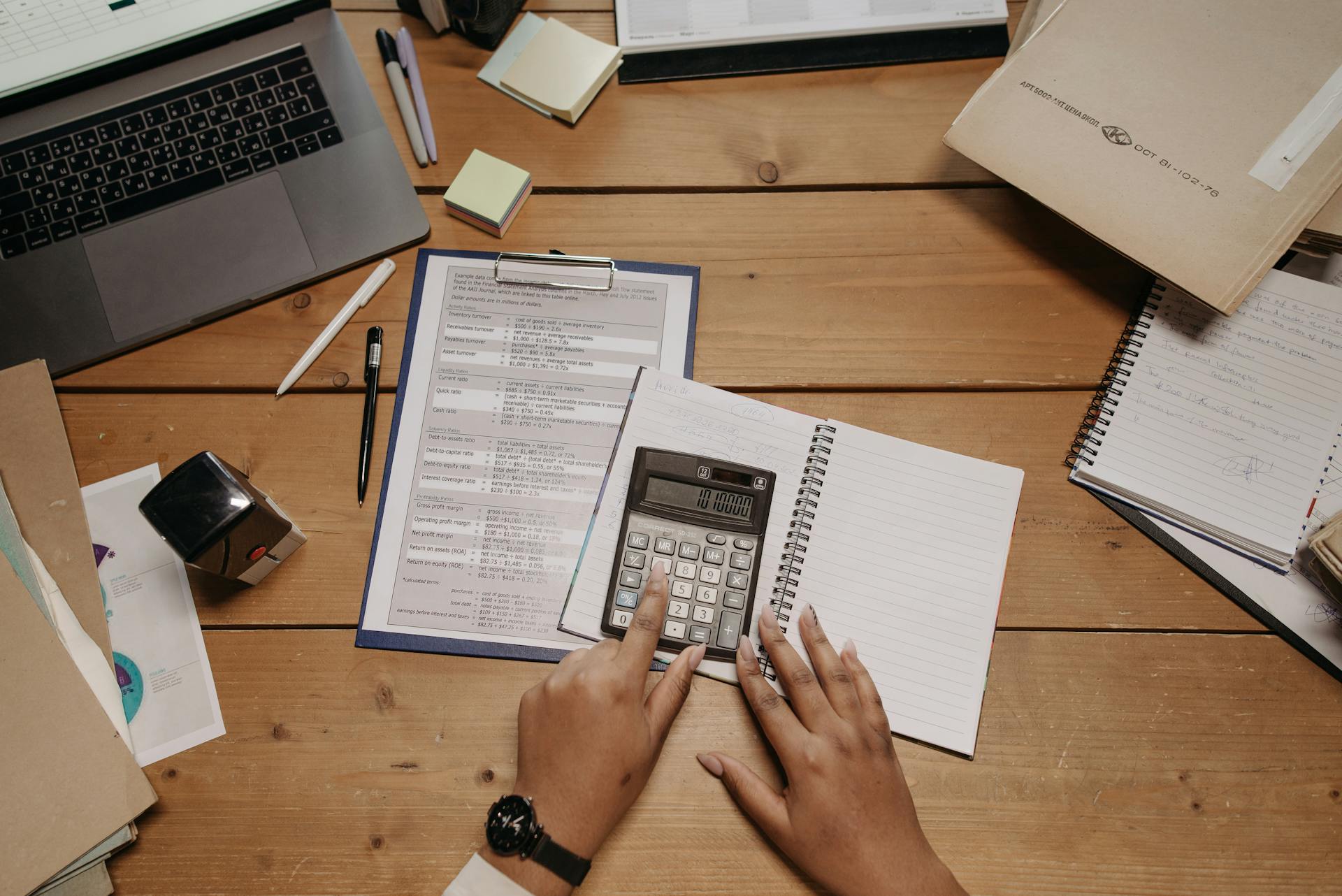 Hands working with calculator and documents on wooden desk in office setting.