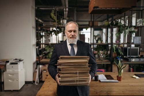 Free Elderly caucasian man with a beard holding piles of paperwork in a modern office environment. Stock Photo
