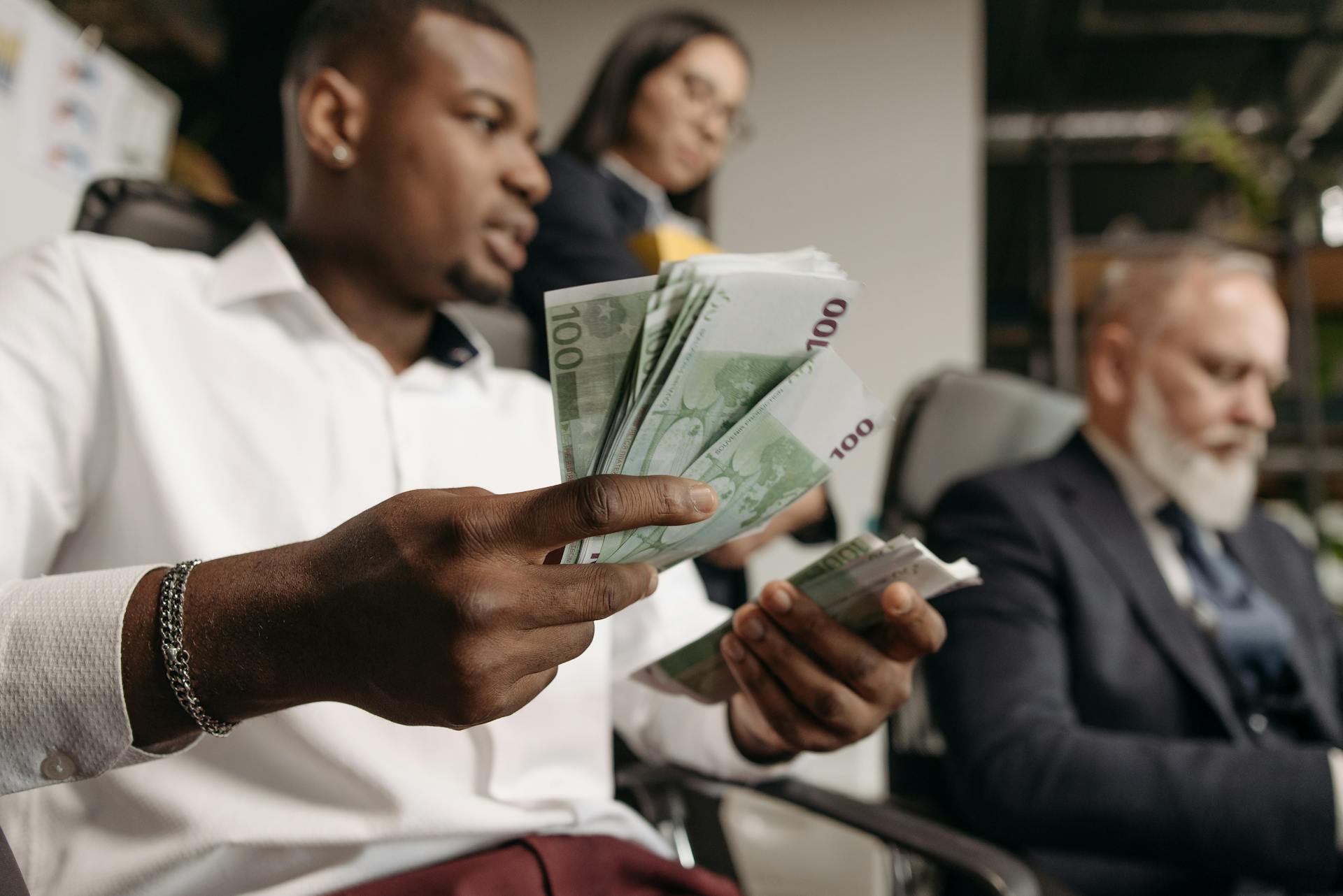 Three professionals in business attire counting cash in a modern office setting.