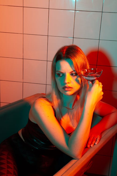 Free Thoughtful young female in stylish black outfit looking away while sitting in bathtub with glass with alcohol drink near tile wall in dark bathroom with neon red and orange lights Stock Photo