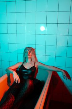 Young slender woman in stylish black clothes lying in bath with glass of alcohol cocktail in dark bathroom near tile wall with neon blue and red lights