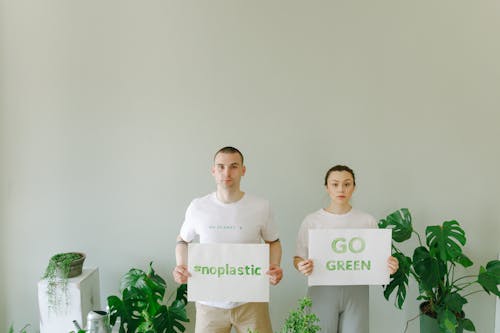 Man and Woman in White Shirt Holding White Banner with Message While Standing Between Green Plants