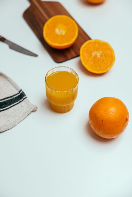 A refreshing vertical shot of fresh orange juice with whole and halved oranges on a white surface, ideal for healthy living concepts.