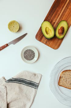 Avocado, bread, lemon, and knife on a cutting board with white background.