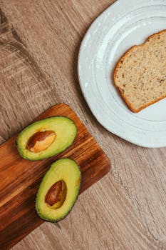 Top view of sliced avocado on a wooden board with a slice of bread on a plate.