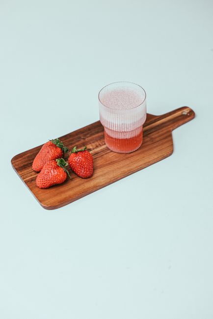 A tall glass of bright red strawberry juice on a wooden board, surrounded by fresh whole and halved strawberries with a few mint leaves.
