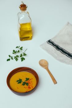 A bowl of pumpkin soup garnished with parsley, next to olive oil and a wooden spoon on a minimalist table.