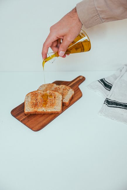 Hand pouring olive oil on wheat toast slices on wooden board.
