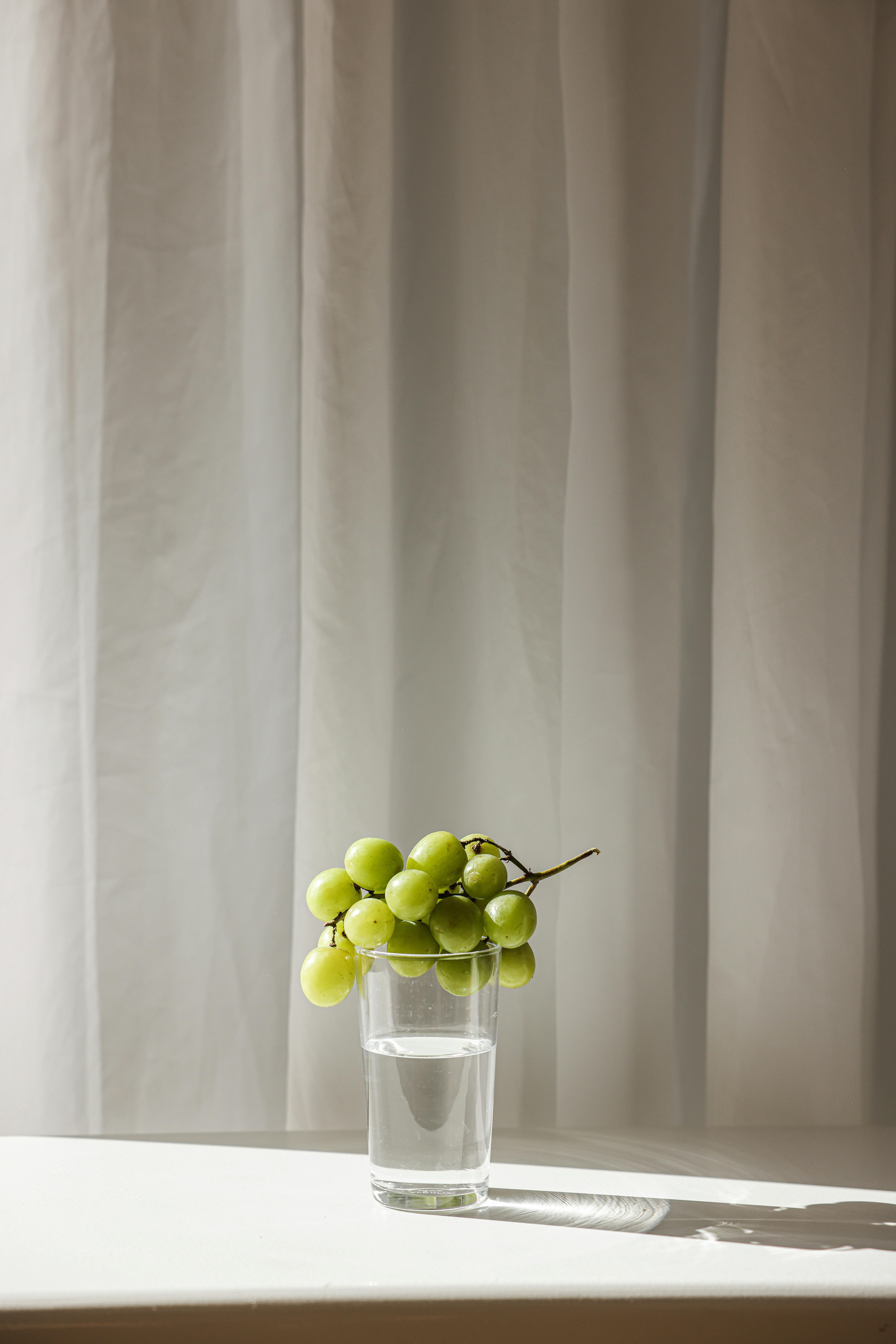 A serene still life featuring green grapes on a glass of water against a curtain backdrop.