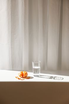 A peeled orange with a glass of water on a sunlit table, creating a minimalist still life scene.