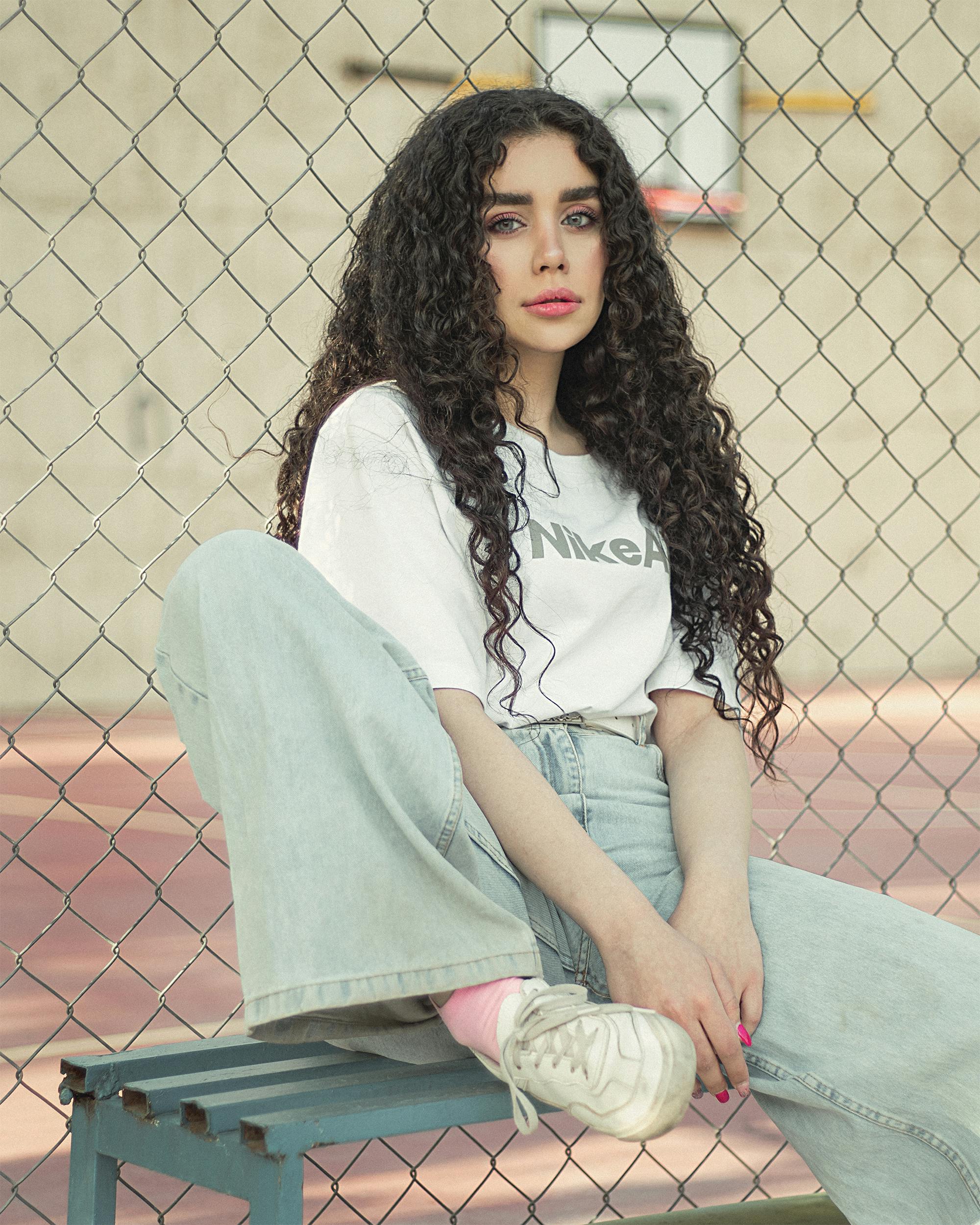 Woman with curly hair and casual denim attire sitting outdoors against a fence.