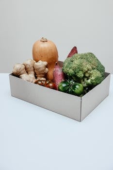 A cardboard box filled with fresh organic vegetables including broccoli, ginger, and squash.