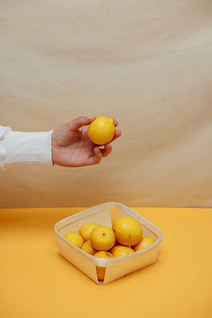 A hand holds a lemon above a basket filled with lemons on a yellow surface, set against a plain backdrop.