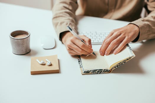 Person writing in a notebook on a desk with coffee and AirPods nearby.