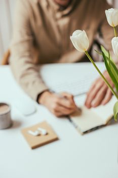 A blurred view of a person writing in a notebook at a minimalist office desk with white tulips.