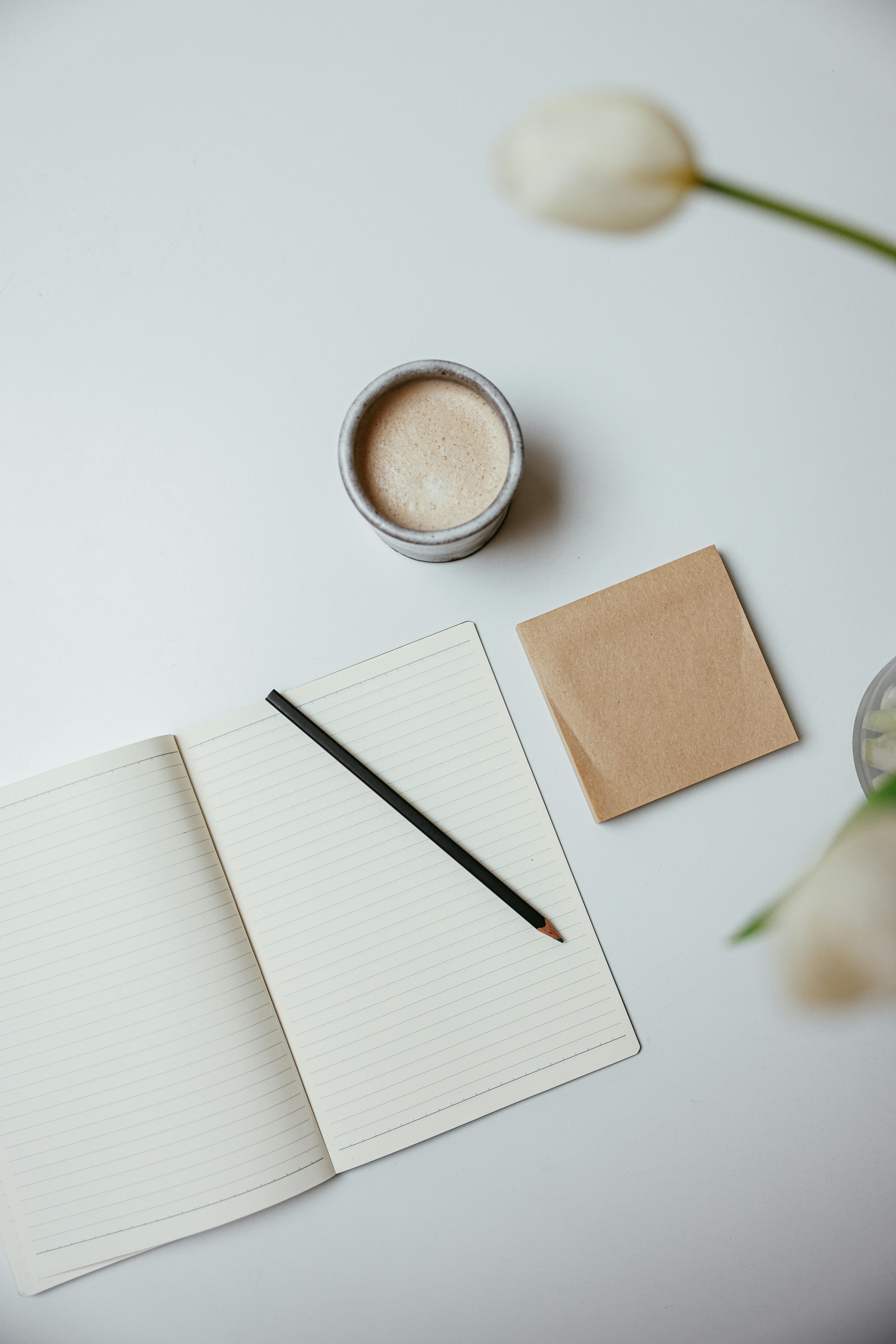 A top view of a minimalist workspace with a notebook, pencil, coffee, and flowers.