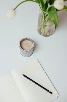 Minimalist setup with a coffee cup, open notebook, and tulip vase on a white desk.