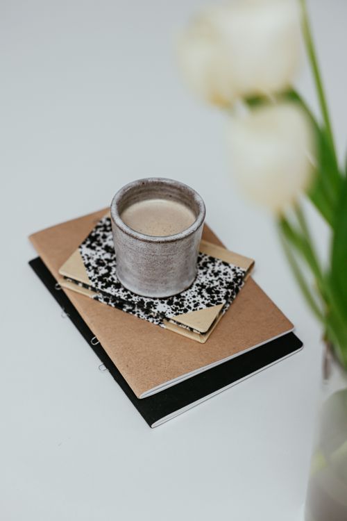 Free A rustic coffee cup sits atop a pile of notebooks with blurred flowers. Stock Photo
