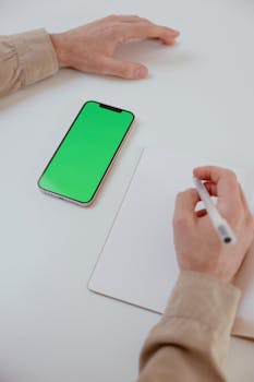 Close-up of hands writing with a smartphone displaying a green screen on a white table.