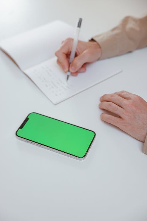 Free Close-Up of a Person Using a Smartphone while Writing on a Notebook Stock Photo