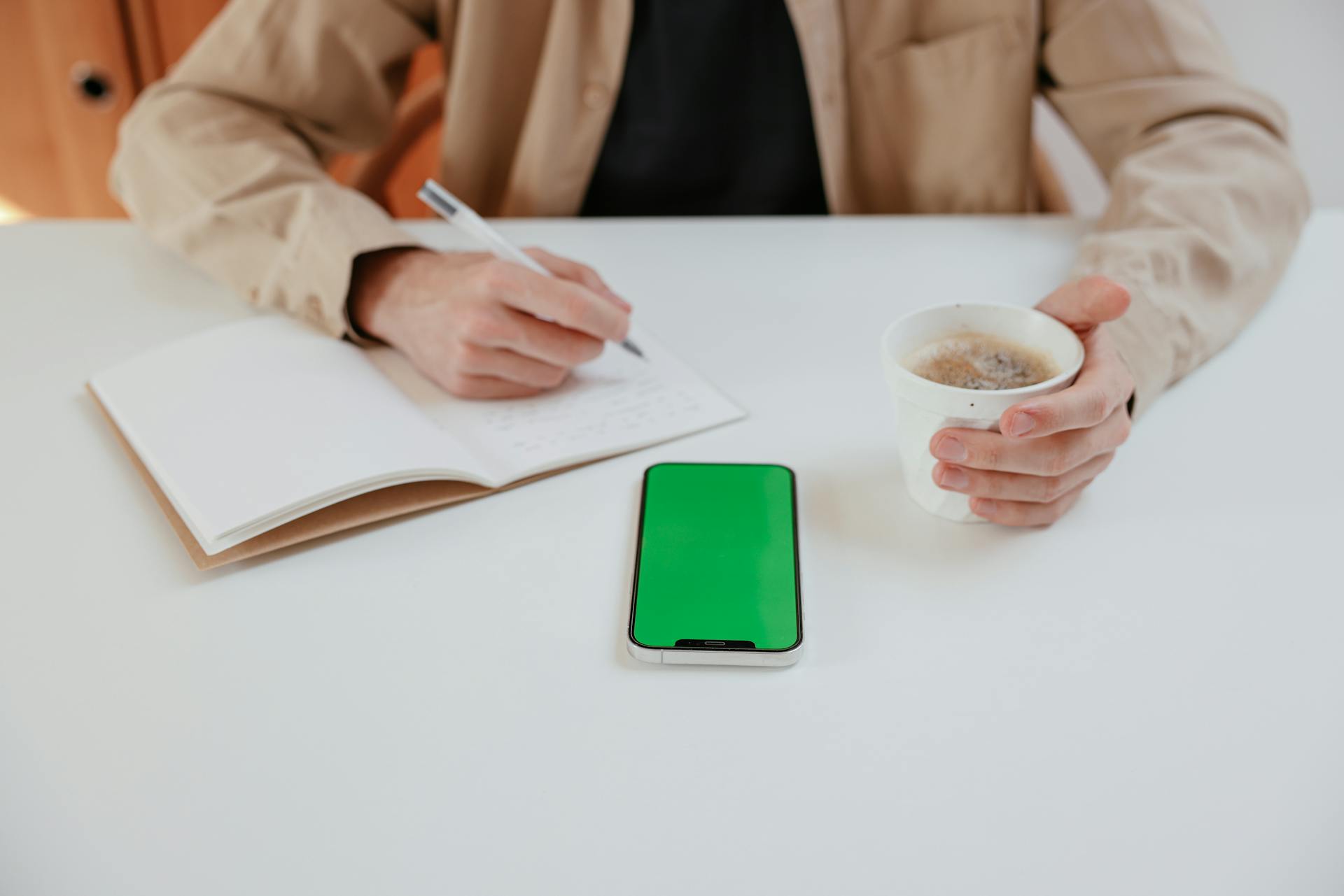 Person Writing on Notebook while Holding a Cup of Coffee