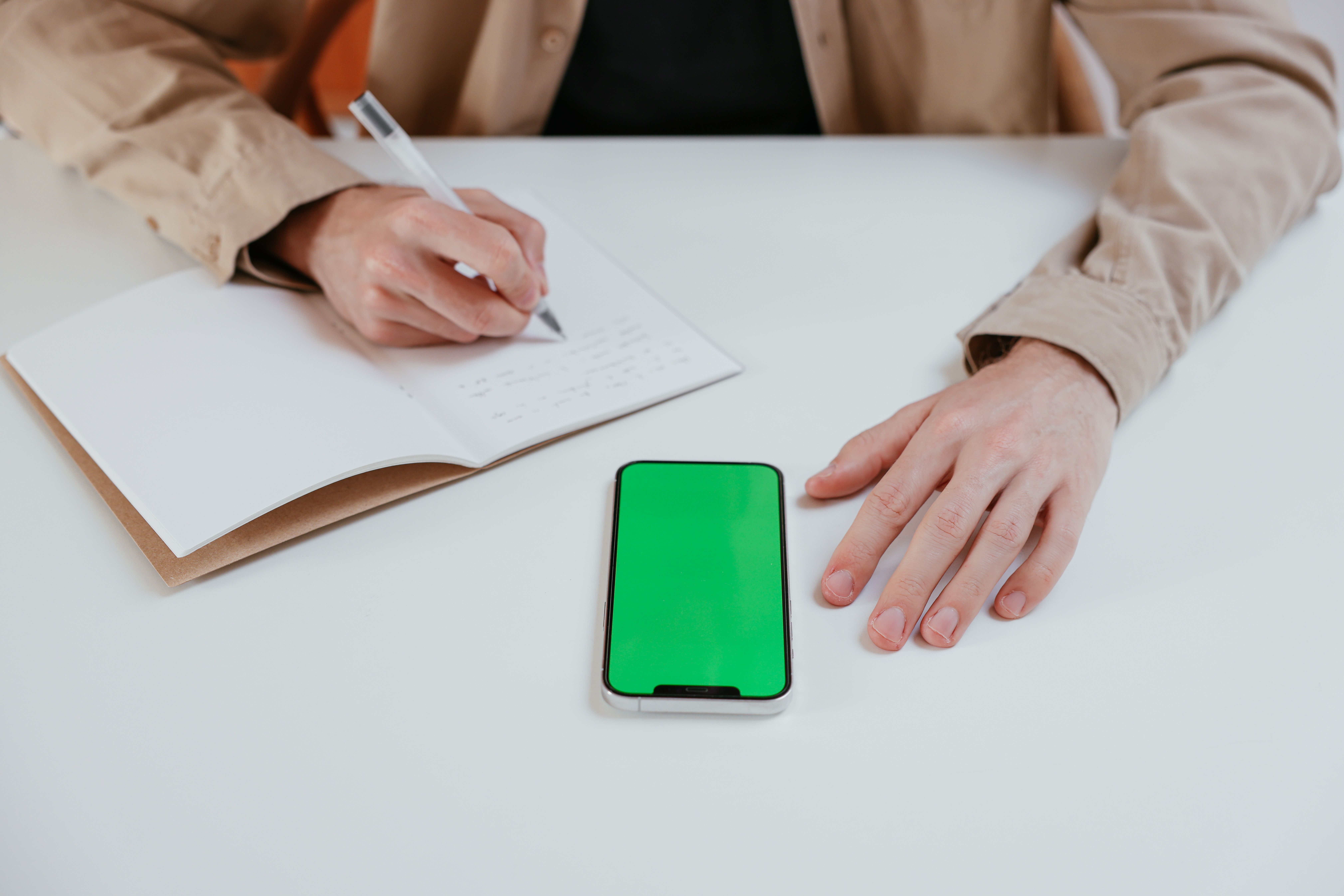 Hands writing in a notebook with a smartphone on a white desk, showcasing productivity.