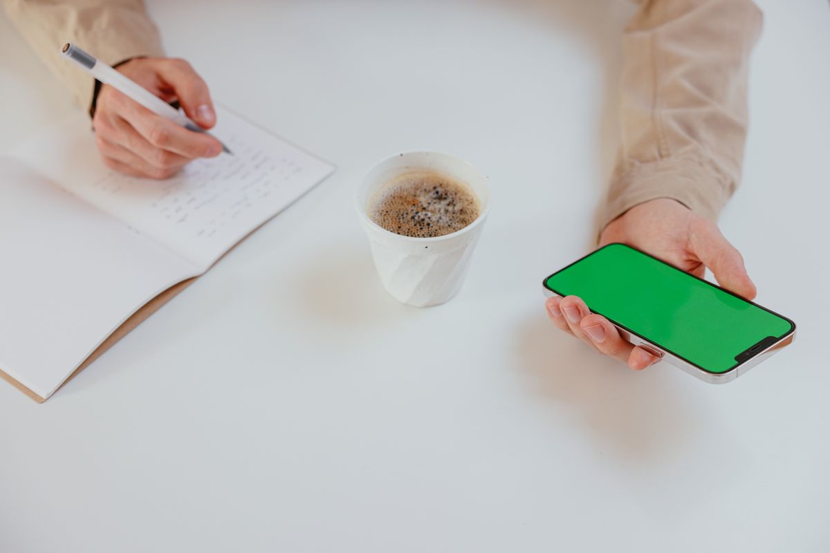 Elinde akıllı telefon tutan kişi ekrana bakıyor, masada kahve fincanı var