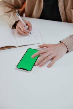 An adult writing in a notebook while touching a smartphone with a green screen on a white table.