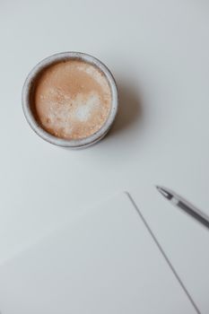 Aerial view of a coffee cup on a white table with a notebook and pen.