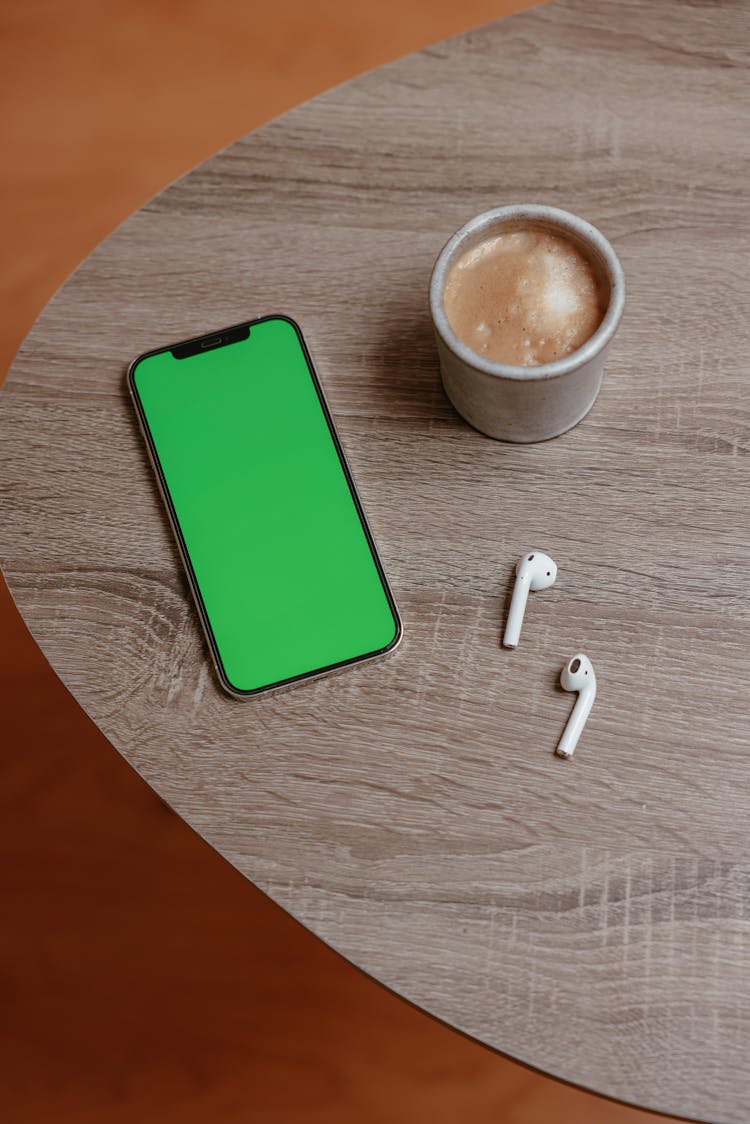 Smartphone With Green Screen And Earbuds Beside A Cup Of Coffee On Wooden Table