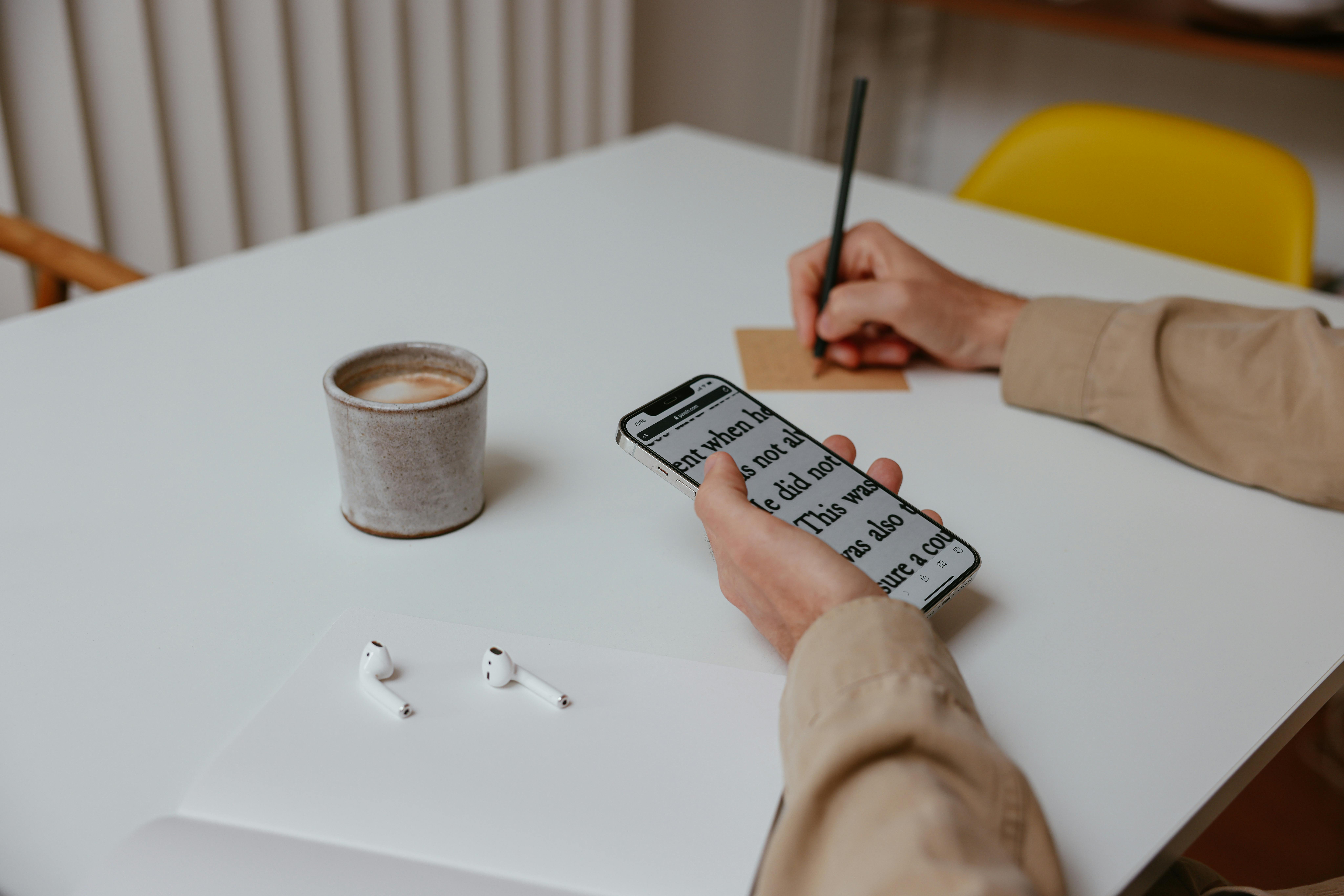 A minimal workspace featuring a cup of coffee, smartphone, earpods, and writing on a white table.