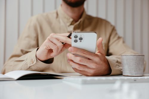 Free Adult man using smartphone with notebook and cup on desk, wearing casual shirt. Stock Photo