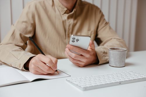 Free Person writing notes while holding a smartphone at a desk, showcasing multitasking. Stock Photo