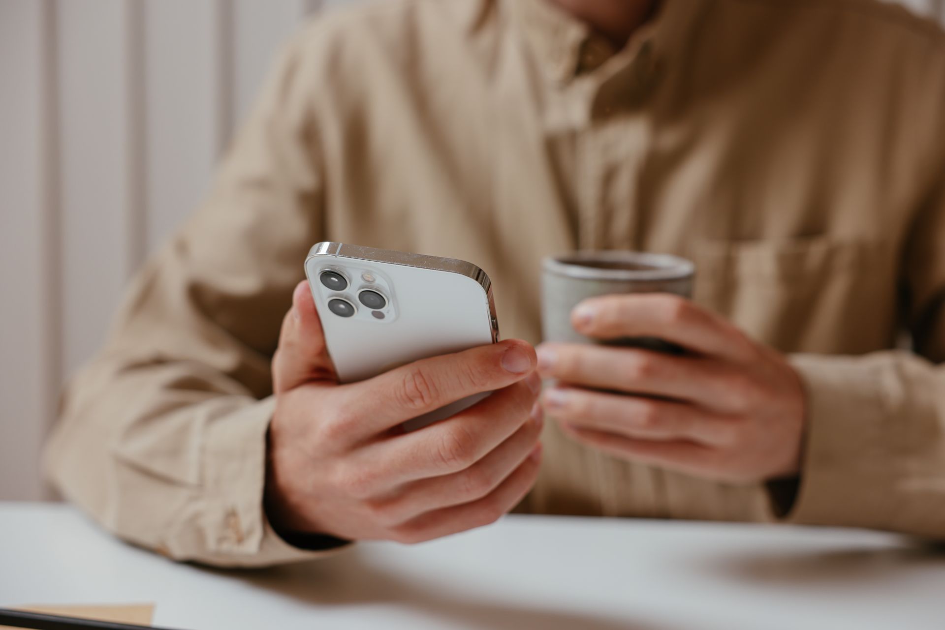 Close-up of a person's hands holding a mobile phone and coffee cup indoors.