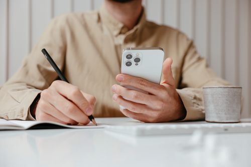 Free A man writes in a notebook while holding a smartphone at an office desk. Stock Photo