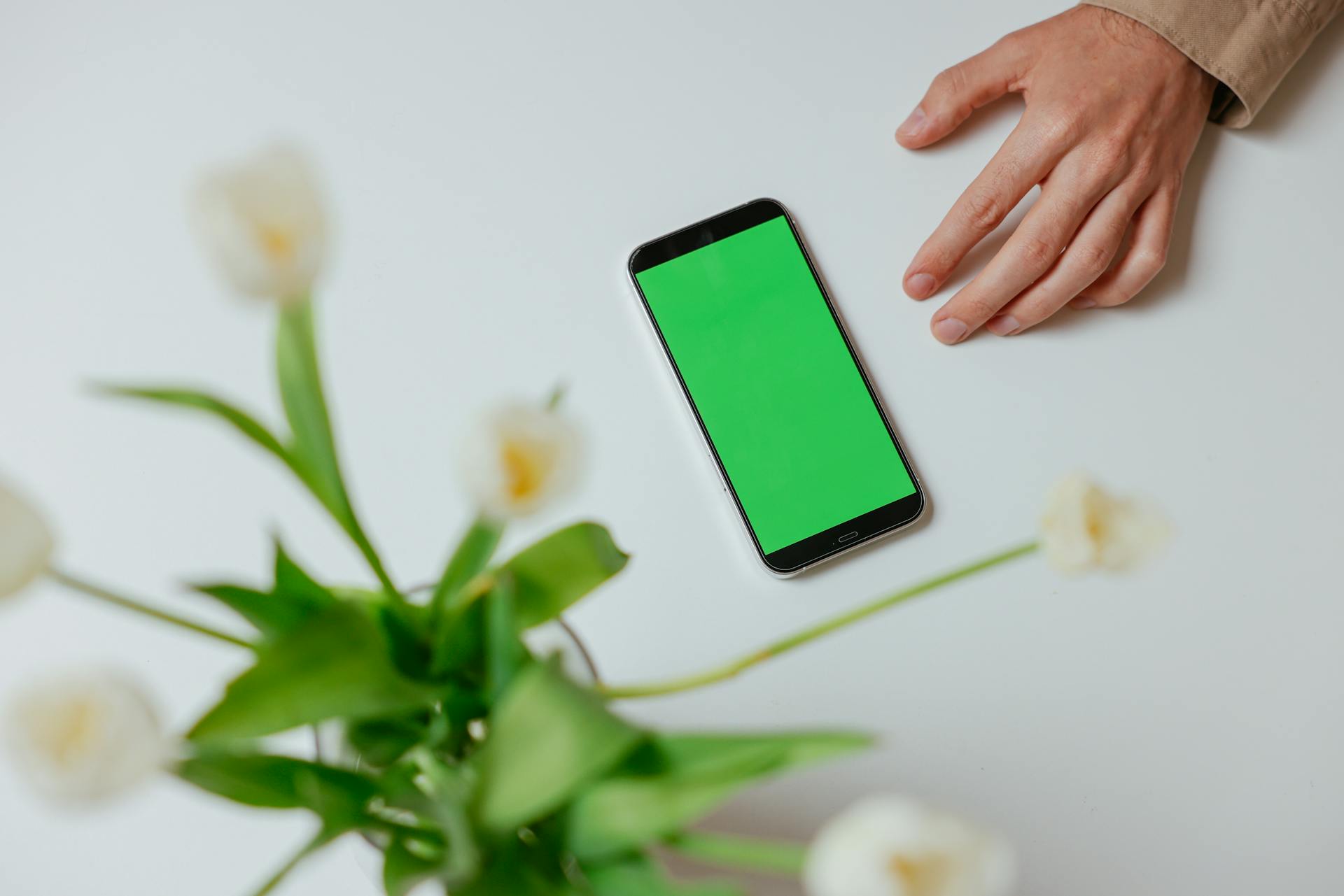 A smartphone with a green screen lies on a table beside a hand and flowers, perfect for design mockups.