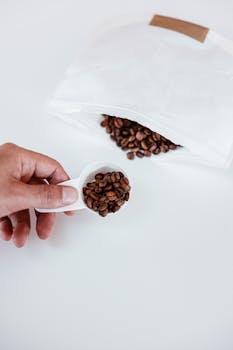 A close-up of a hand holding a measuring spoon filled with coffee beans next to a white bag.