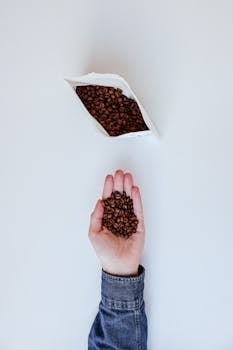 Top view of a hand holding coffee beans, with a white bag on a clean white surface.