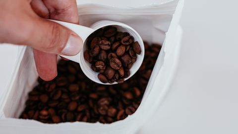 A hand holding a measuring spoon with coffee beans over an open bag on a white background.