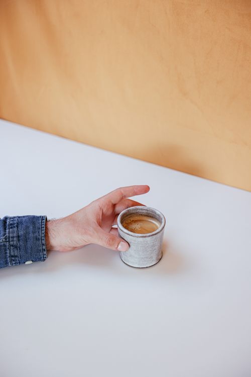 Free Minimalist image of a hand in denim holding a ceramic coffee cup on a white table. Stock Photo