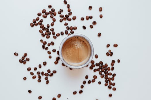 A steaming cup of coffee surrounded by scattered roasted coffee beans on a white surface.
