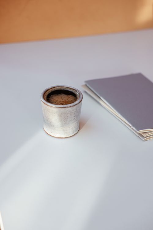 Free Warm cup of coffee next to a closed notebook on a minimalist white table setting. Stock Photo