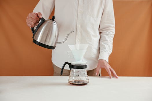 A person brewing pour over coffee using a dripper and kettle in a minimalistic setting.