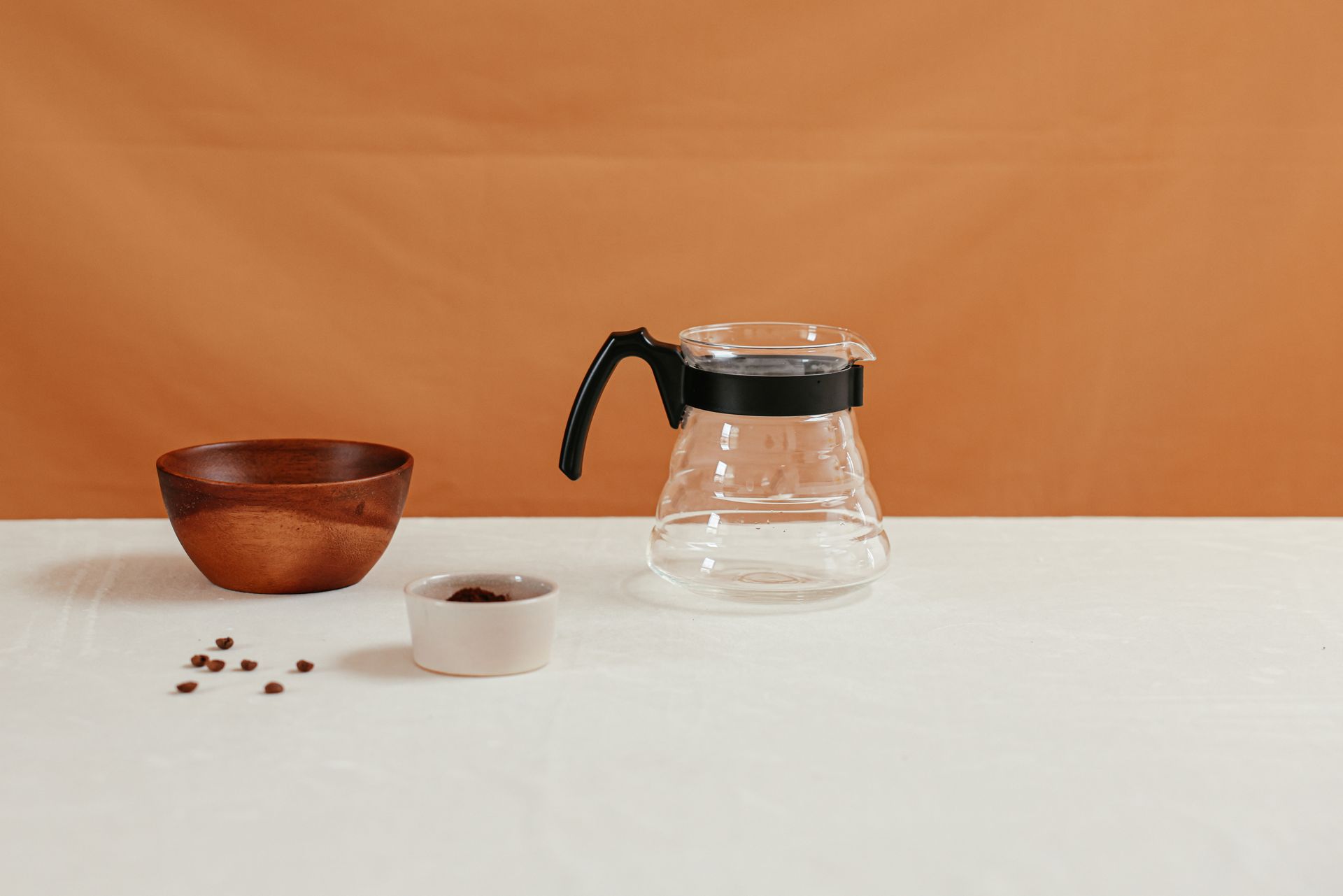 A minimalist coffee setup featuring a glass coffee maker with a wooden and ceramic bowl on a white surface against an orange background.