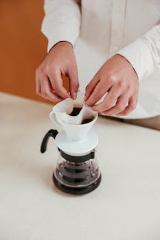 Close-up of hands making pour over coffee using a dripper and filter in a warm setting.
