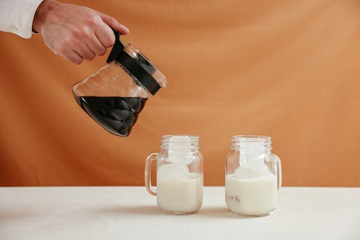 A close-up of a hand pouring coffee from a server into glasses of iced milk.